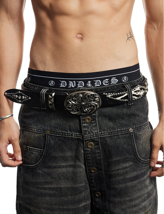 A close-up of a man's lower torso wearing a black studded swag belt with a large ornate silver buckle, styled with distressed black swag jeans. The belt features the brand name 'DND' in white on the waistband.