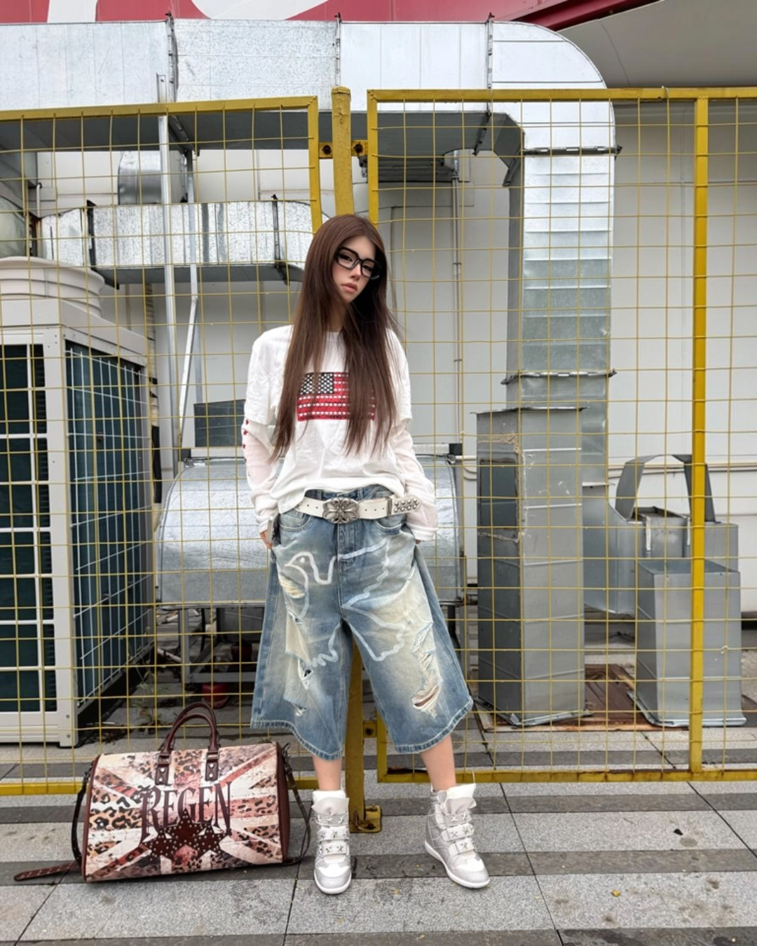 Person wearing a white sweatshirt with an American flag design and ripped jeans, standing next to a patterned bag in an urban setting.